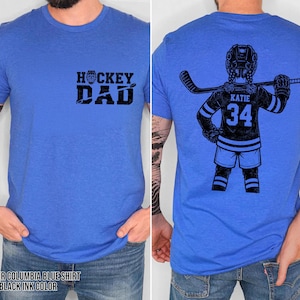 Pode incluir: Camiseta azul Columbia mesclada com tinta preta. A frente exibe "HOCKEY DAD" em negrito. A parte de tr&aacute;s apresenta um gr&aacute;fico de jogador de h&oacute;quei com o nome "KATIE" e o n&uacute;mero "34".