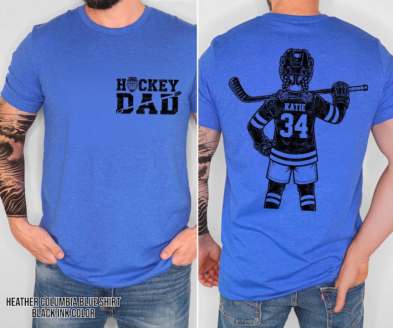 Pode incluir: Camiseta azul Columbia mesclada com tinta preta. A frente exibe "HOCKEY DAD" em negrito. A parte de tr&aacute;s apresenta um gr&aacute;fico de jogador de h&oacute;quei com o nome "KATIE" e o n&uacute;mero "34".