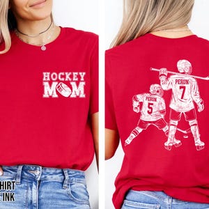 May include: Red t-shirt with white graphic text and design. The front of the shirt says "HOCKEY MOM" with a hockey puck graphic. The back of the shirt features two hockey players with the text "PERUN" and the number "7" and "5".