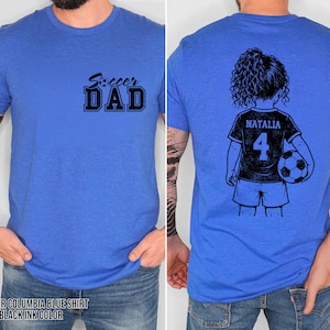 May include: Heather Columbia blue t-shirt with the text "Soccer Dad" in black on the front. The back features a black and white illustration of a girl holding a soccer ball, with the name "Natalia" and the number "4" on her jersey.