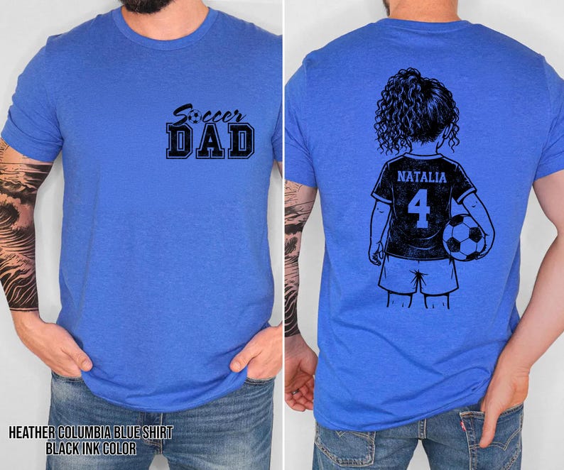 May include: Heather Columbia blue t-shirt with the text "Soccer Dad" in black on the front. The back features a black and white illustration of a girl holding a soccer ball, with the name "Natalia" and the number "4" on her jersey.