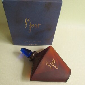 May include: A brown, geometric-shaped perfume bottle with a blue stopper and the text "le jour" in gold script. The bottle is next to a dark blue box with the same text and "Eau de Toilette". The bottle contains 1.6 fl oz.