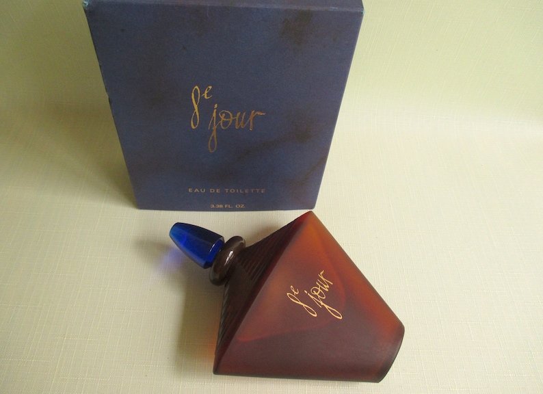 May include: A brown, geometric-shaped perfume bottle with a blue stopper and the text "le jour" in gold script. The bottle is next to a dark blue box with the same text and "Eau de Toilette". The bottle contains 1.6 fl oz.