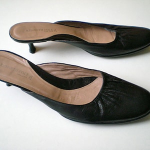May include: A pair of black leather heeled mules with a low heel. The shoes have a rounded toe and a slightly pointed back. The brand name "Kenneth Cole II" is printed on the insole.