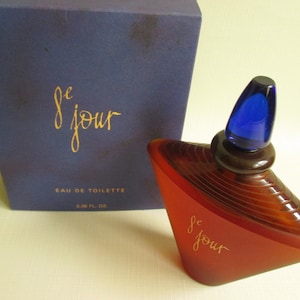 May include: A perfume bottle and box set. The bottle is a unique shape, with a red-brown body and a blue stopper. The box is dark blue with gold lettering that reads "le jour" and "Eau de Toilette". The bottle contains 3.38 fl oz.