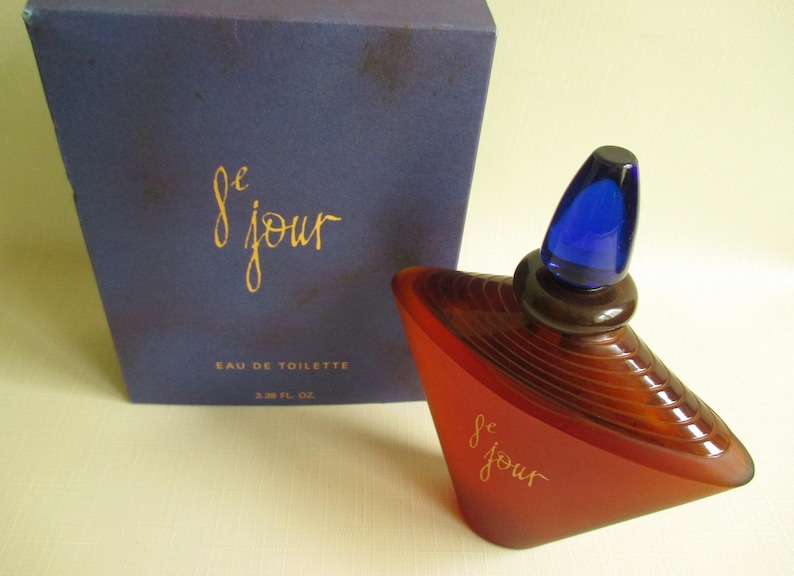 May include: A perfume bottle and box set. The bottle is a unique shape, with a red-brown body and a blue stopper. The box is dark blue with gold lettering that reads "le jour" and "Eau de Toilette". The bottle contains 3.38 fl oz.