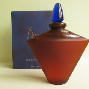May include: A geometric-shaped perfume bottle with a dark red frosted finish and a blue stopper. The bottle is next to a navy blue box with gold script. The bottle is labeled "Eau de Toilette" and is 3.38 fl oz.