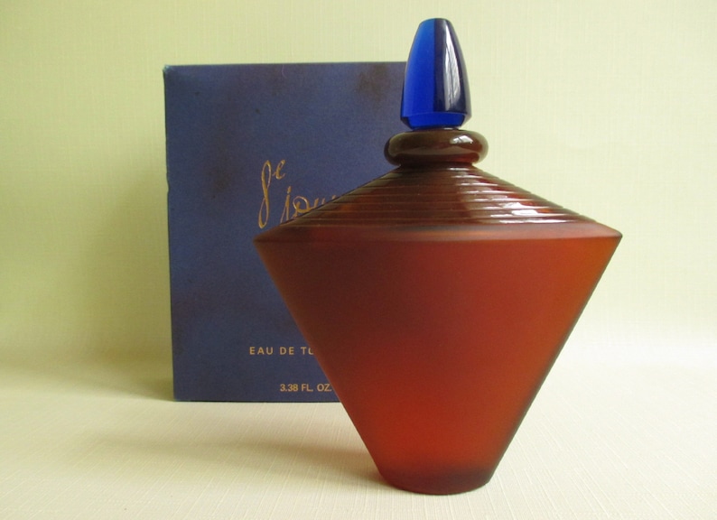 May include: A geometric-shaped perfume bottle with a dark red frosted finish and a blue stopper. The bottle is next to a navy blue box with gold script. The bottle is labeled "Eau de Toilette" and is 3.38 fl oz.