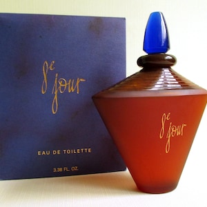 以下が含まれることがあります： 香水瓶と箱のセット。ボトルは赤茶色で、青いストッパーと金色の筆記体で「le jour」と書かれています。箱は濃い青色で、同じ金色の筆記体と「Eau de Toilette」の文字が書かれています。