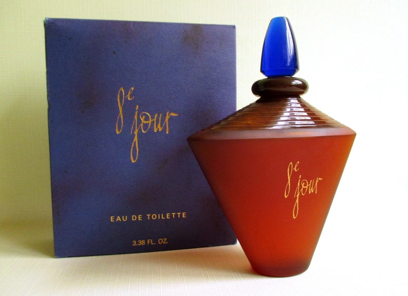 May include: A perfume bottle and box set. The bottle is a reddish-brown color with a blue stopper and the words "le jour" in gold script. The box is dark blue with the same gold script and the words "Eau de Toilette".