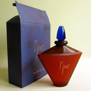 May include: A perfume bottle and box set. The bottle is a reddish-brown color with a blue cap and the words "le jour" in gold script. The box is dark blue with the same gold script.