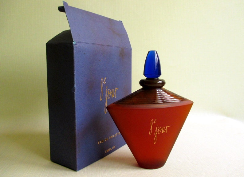 May include: A perfume bottle and box set. The bottle is a reddish-brown color with a blue cap and the words "le jour" in gold script. The box is dark blue with the same gold script.