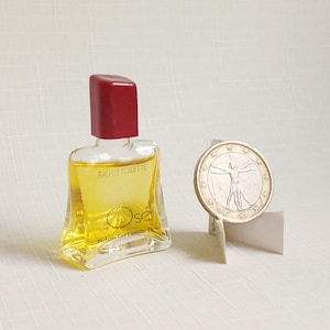 Può includere: Una piccola bottiglia di vetro trasparente di Ose Eau de Toilette con tappo rosso. La bottiglia contiene liquido giallo. Una moneta con una figura umana è appoggiata su un piccolo supporto bianco.