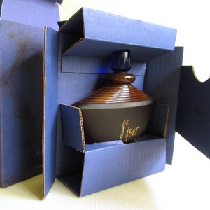 May include: A dark brown perfume bottle with a brown and gold design, and a blue stopper, is nestled inside a blue cardboard box. The bottle has the word "le jour" in gold script. The box is open, revealing the bottle.