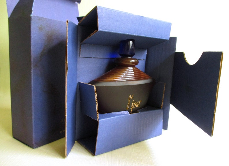 May include: A dark brown perfume bottle with a brown and gold design, and a blue stopper, is nestled inside a blue cardboard box. The bottle has the word "le jour" in gold script. The box is open, revealing the bottle.