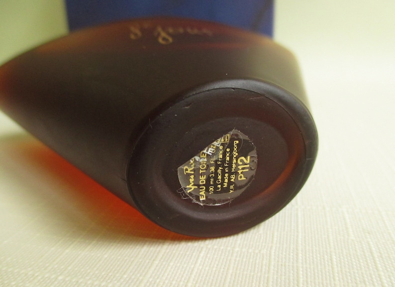 May include: A brown perfume bottle with a rounded shape. The bottle has a label with text including "EAU DE TOILETTE" and "100 ml 3.38 FL. OZ.". The bottle is made in France.