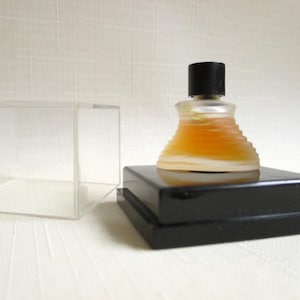 May include: A small, vintage perfume bottle with a black cap, filled with amber liquid, sits on a black, rectangular stand. The bottle has a unique, tiered design. A clear, rectangular box is to the left.