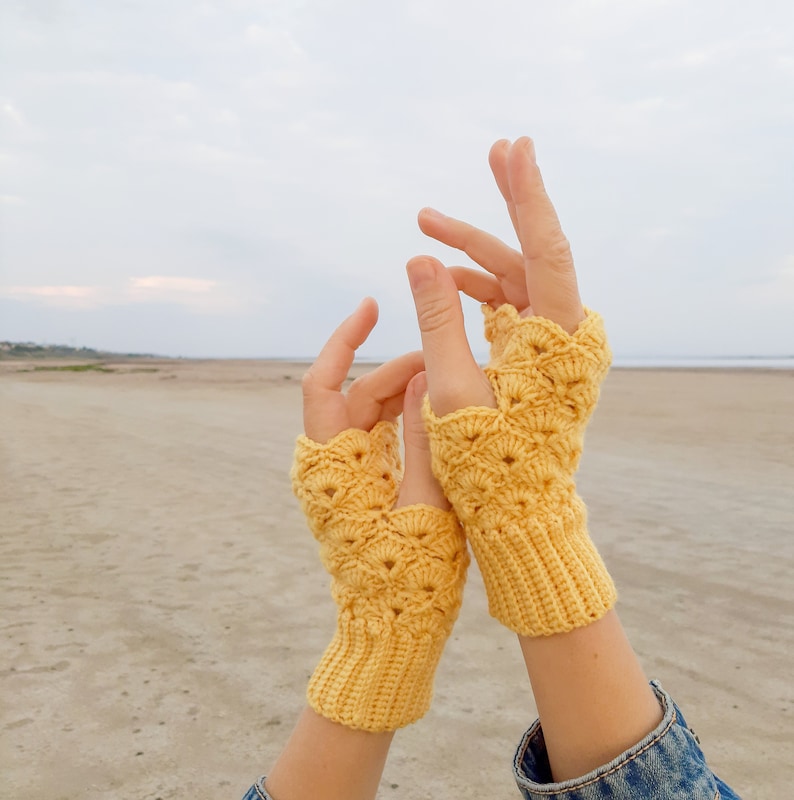 May include: A pair of yellow crocheted fingerless gloves with a lacy pattern. The gloves are worn by a person with their hands raised in the air.