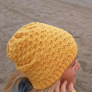Op de afbeelding: Een gele gebreide beanie met een textuurpatroon. De beanie wordt gedragen door een persoon met blond haar.