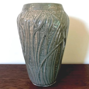 May include: A sage green ceramic vase with a glossy finish and a textured, raised leaf-like pattern across its surface. The vase features a tapered cylindrical shape with a smooth rim. A copper penny is placed next to the base for scale.