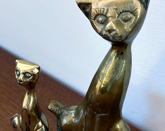 May include: Two brass cat figurines. One is small and seated, the other is larger and upright. Both have detailed facial features and a polished finish. The cats are displayed on a wooden surface.