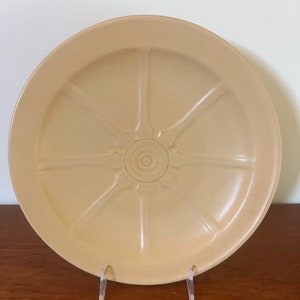 Puede incluir: Un plato grande de cerámica beige con un diseño en forma de rueda. El plato tiene un centro elevado con un patrón circular y seis radios que irradian hacia afuera.