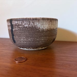 May include: A small, handmade ceramic bowl with a textured surface. The bowl features a brown and white color scheme with a black vertical stripe. A penny is next to the bowl for size reference.
