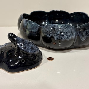 May include: A dark blue ceramic serving bowl with a matching lid. The bowl has a unique, scalloped shape and a glossy finish with white and blue accents. The lid features a decorative handle. A penny is next to the lid.