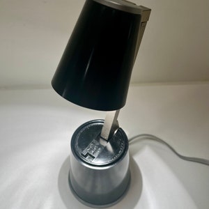May include: A black desk lamp with a conical shade and a silver-colored base. The lamp has a flexible arm and a round base. The lamp is plugged into a wall outlet with a gray cord.