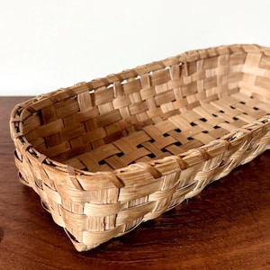 May include: A rectangular woven basket made of light brown natural splints with a simple over-under weave pattern. The basket features a reinforced rim and a flat base. A single copper-colored penny is placed nearby for scale reference.