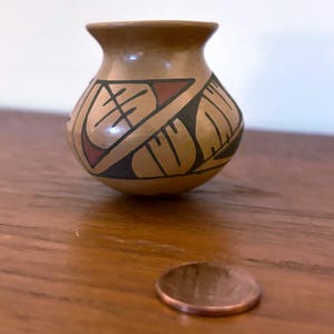 May include: Small, brown ceramic vase with geometric designs in black and red. The vase has a rounded body and a narrow neck. A penny is next to the vase for size comparison. The vase is on a wooden surface.