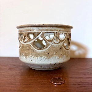 May include: A small, handcrafted ceramic pot with a decorative openwork design around the top. The pot has a neutral color palette of beige and brown, with a slightly textured surface. A penny is next to the pot.