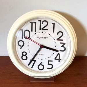 May include: A round wall clock with a cream-colored frame and a white face. The clock features large black Arabic numerals, black hour and minute hands, and a thin red second hand. The text Ingraham, QUARTZ, and USA are printed on the face.