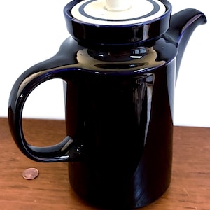 May include: A glossy, dark blue ceramic teapot with a matching lid. The lid has a white knob and a blue and white ring design. The teapot has a curved handle and a spout. A penny is next to the teapot.