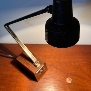 May include: A black desk lamp with a textured finish, featuring a brass arm and a wooden base. The lamp is positioned on a wooden surface. The lamp's arm is adjustable.