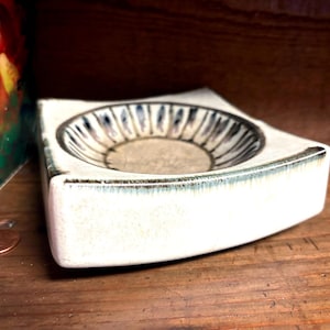 May include: A square ceramic dish with a white exterior and a recessed circular center featuring a dark blue and white radial pattern. The edges of the dish show a subtle dark blue glaze accent, and the overall form has slightly curved sides.