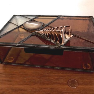 May include: A stained glass box with a hinged lid, featuring a geometric design in shades of brown and amber. A decorative seashell and a polished stone adorn the top. The box is on a wooden surface, with a penny for scale.