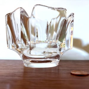 May include: A clear, faceted glass votive candle holder with a unique, scalloped rim. The clear glass reflects light, creating a sparkling effect. The candle holder sits on a wooden surface, with a penny nearby for scale.