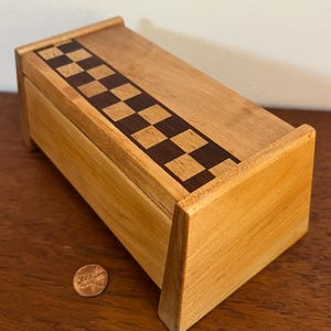 May include: A small wooden box with a checkerboard inlay on the lid. The box is made of light wood and has a natural finish. A penny is placed next to the box for scale.