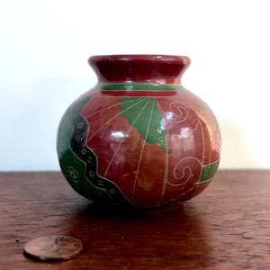 May include: A small, round, hand-painted vase with a dark red base and a green and red design. The vase has a dark red rim and is sitting on a wooden surface. A penny is next to the vase.