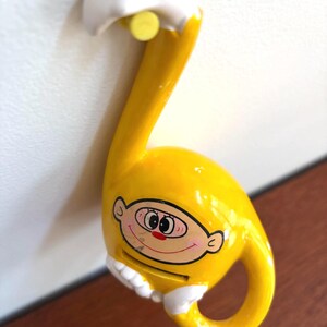 May include: A yellow ceramic money box in the shape of a musical note, featuring a cartoon face with a red nose and smiling mouth. It has a coin slot and a handle. The money box is attached to a white surface.