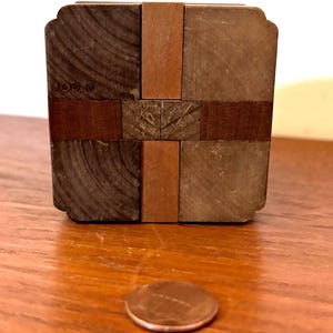 May include: A wooden puzzle box with a cross design, featuring alternating dark and light brown wood pieces. The word "JAPAN" is visible on one side. A penny is in the foreground.