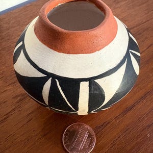 Peut inclure: Petit pot rond en céramique peint à la main, avec une large ouverture. Le pot présente un motif de formes géométriques noires et blanches sur fond crème. Le bord est brun rougeâtre. Une pièce de monnaie est à côté du pot pour la référence de taille.