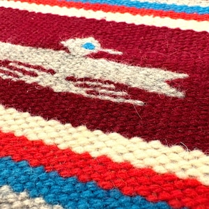 May include: Close-up of a woven textile with a maroon background and a white bird design. The textile features horizontal stripes in red, white, and blue. The bird has a blue eye.