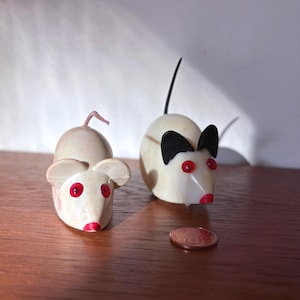 May include: Two small mouse ornaments. One is off-white with red eyes and a red nose, the other is white with black ears, red eyes, and a red nose. A penny is next to the mice on a wooden surface.