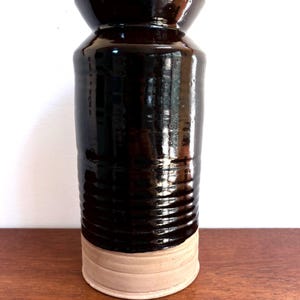 May include: A tall, dark brown ceramic vase with a glossy finish. The vase has a cylindrical body with horizontal ridges and a lighter beige base. The top flares out slightly. A penny is next to the vase.