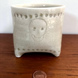 May include: A small, off-white ceramic cup with a skull design. The cup has a square shape with three small feet. The skull is etched into the front of the cup, and the cup has a textured pattern. A penny is next to the cup.