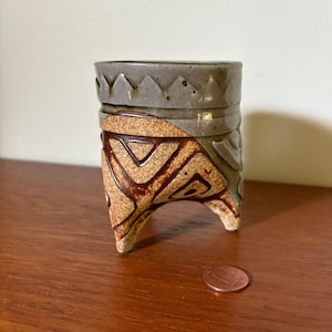 May include: A small, handmade ceramic pot with a unique design. The pot is a muted gray color on top, transitioning to a tan and brown geometric pattern. It stands on three small legs and is approximately 3 inches tall.