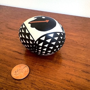 May include: A decorative ceramic ball with a black and white geometric design. The ball features a black cloud shape with a red arrow on a white background. A penny is next to the ball on a wooden surface.