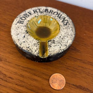 Puede incluir: Un cenicero redondo con el texto "ROBERT BROWN'S". El cenicero tiene una superficie moteada blanquecina con un cuenco central amarillo-marrón. Una moneda está al lado del cenicero.